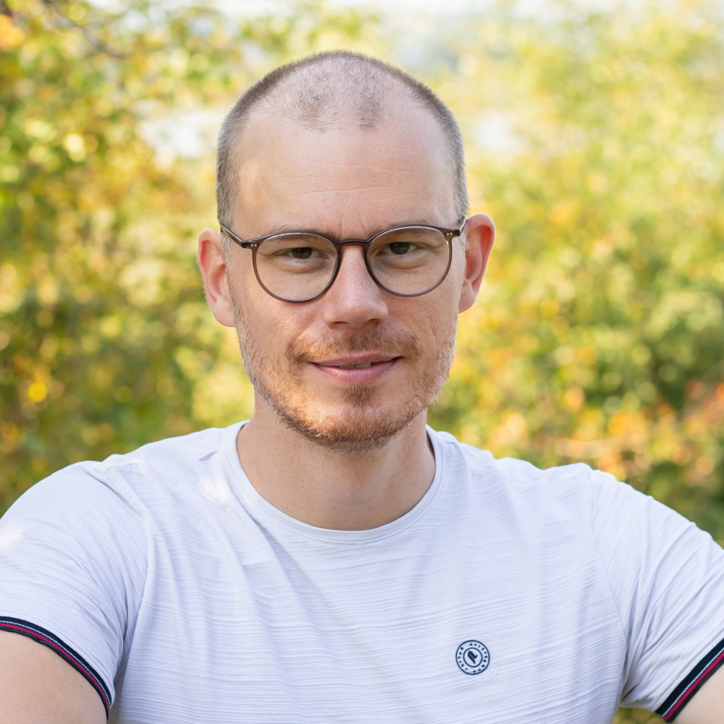 Porträt von Christopher Kamper mit Brille und weißem T-Shirt vor herbstlichem Grün.