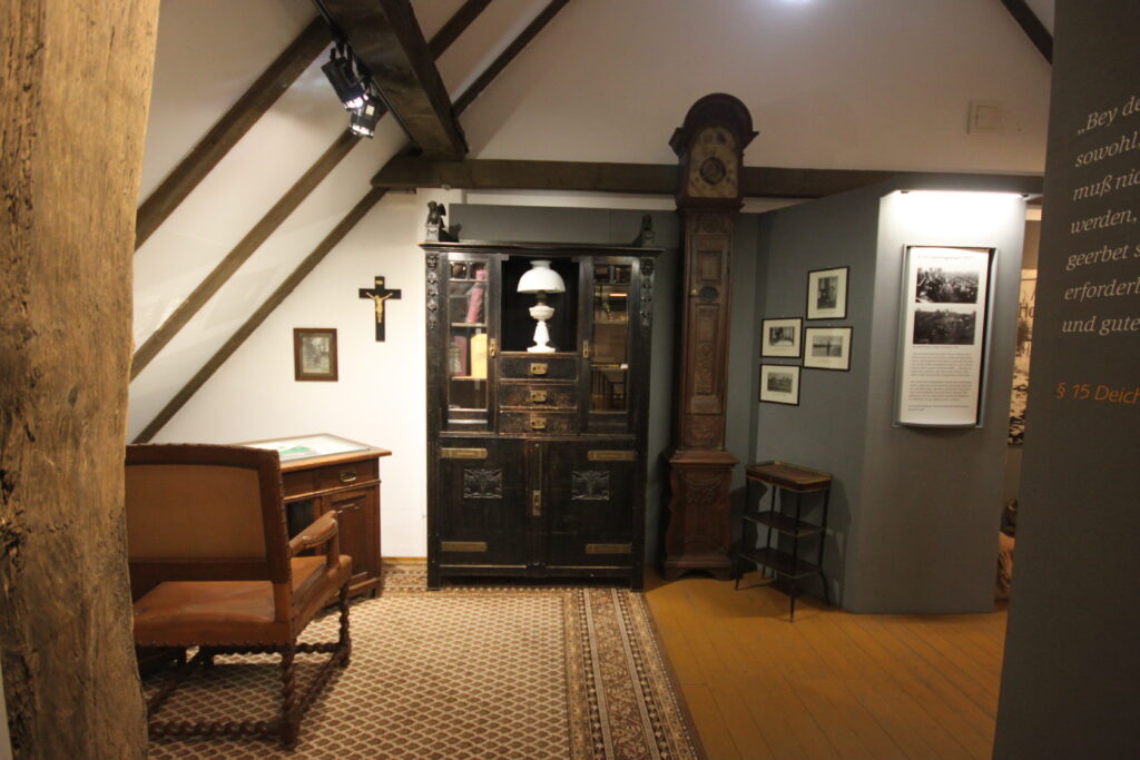 Der Innenraum eines historischen Museums mit einer antiken Standuhr, einem Schreibtisch und verschiedenen Bildern an den Wänden.