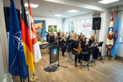 Ein Blasorchester mit mehreren Musikern spielt in einem hellen Raum, umgeben von Flaggen und Emblemen.