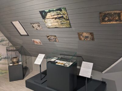 Im Raum sind Vitrinen mit historischen Artefakten und Tafeln mit Bildern und Informationen an der Wand angebracht.