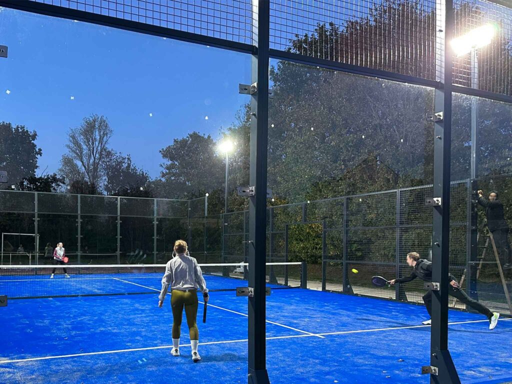 Zwei Spielerinnen und ein Spieler beim Padel-Tennis auf einem beleuchteten Platz in der Dämmerung.