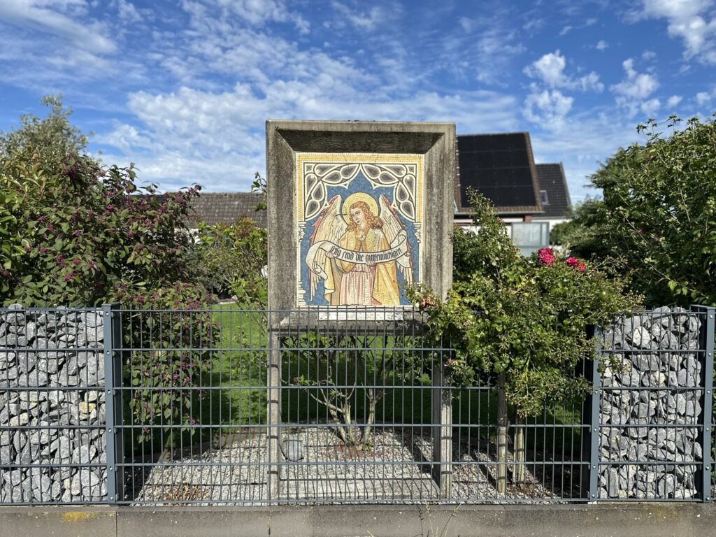 Eine religiöse Mosaikdarstellung eines Engels ist in einen Garten mit Büschen und Himmel im Hintergrund eingefasst.