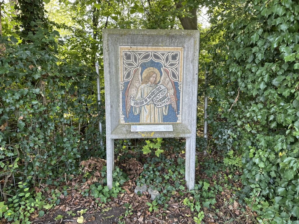 Ein Mosaik eines Engels steht auf einem Betonsockel, umgeben von grünen Sträuchern und abgefallenem Laub.