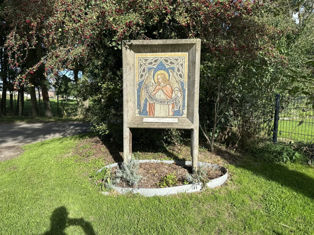 Ein Mosaikbild eines Heiligen in einem Rahmen steht auf einem grünen Rasen umgeben von Blumen und Sträuchern.