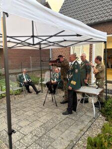 Fünf Männer in Uniformen stehen und sitzen unter einem Pavillon auf einem Gehweg. Ein Mann bedient ein Kamerastativ.
