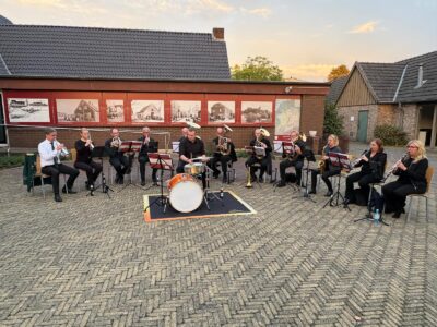 Eine Blaskapelle mit mehreren Musikern spielt im Freien, umgeben von historischen Fotografien an einer Wand.