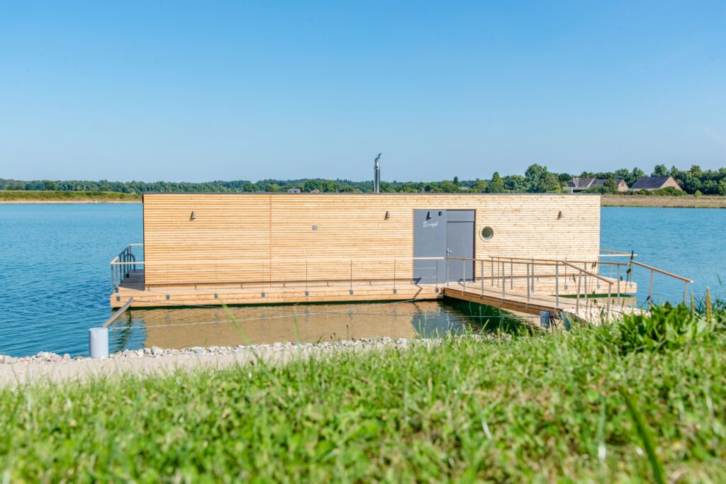Holzhaus auf einer Plattform im Wasser, umgeben von Grüns und einem klaren blauen Himmel.