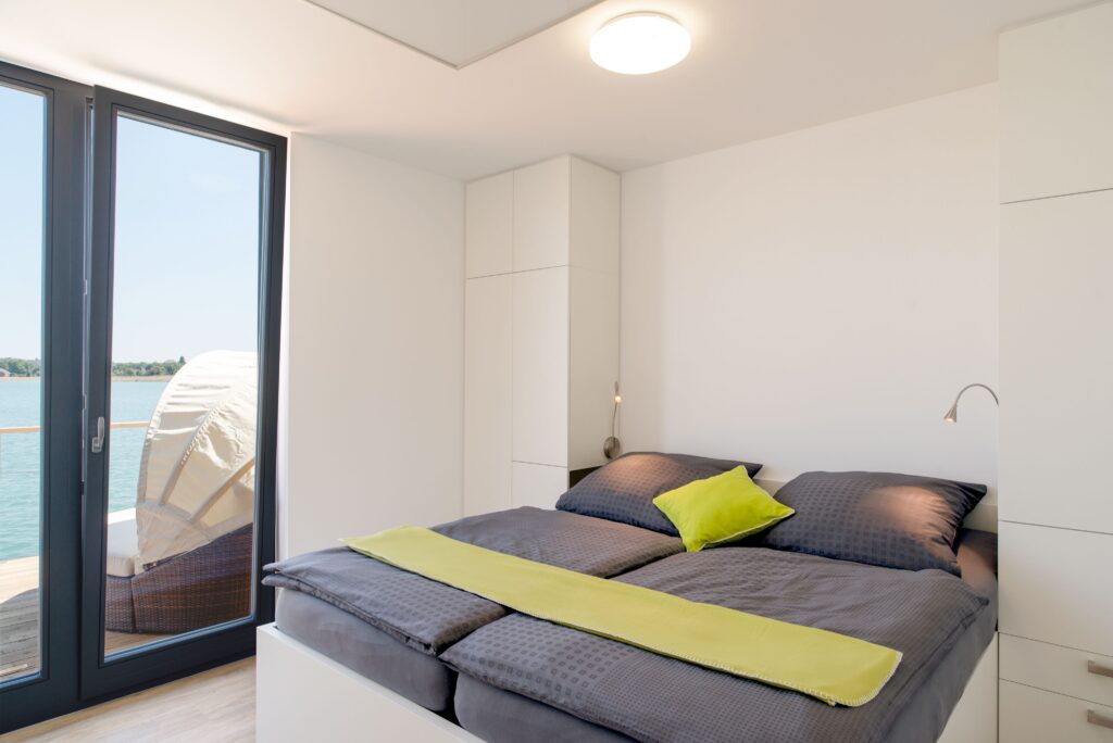Helle, moderne Schlafzimmeransicht mit einem Doppelbett, einem Kissen in Limettengrün und Fensterblick auf Wasser.