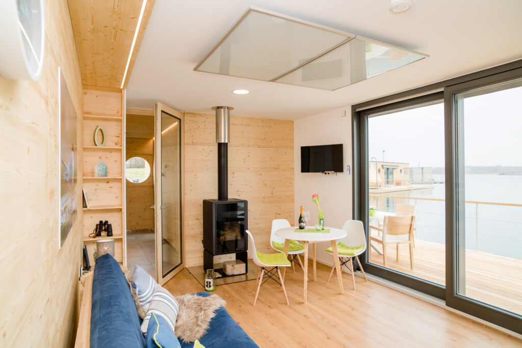 Helles Wohnzimmer mit Holzverkleidung, Tisch und Stühlen, Kamin und großen Fenstern mit Blick auf das Wasser.