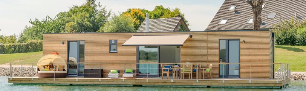 Modernes Holzhaus auf dem Wasser mit Terrasse, Liegestühlen und Sonnenschutz, umgeben von grüner Landschaft und Bäumen.