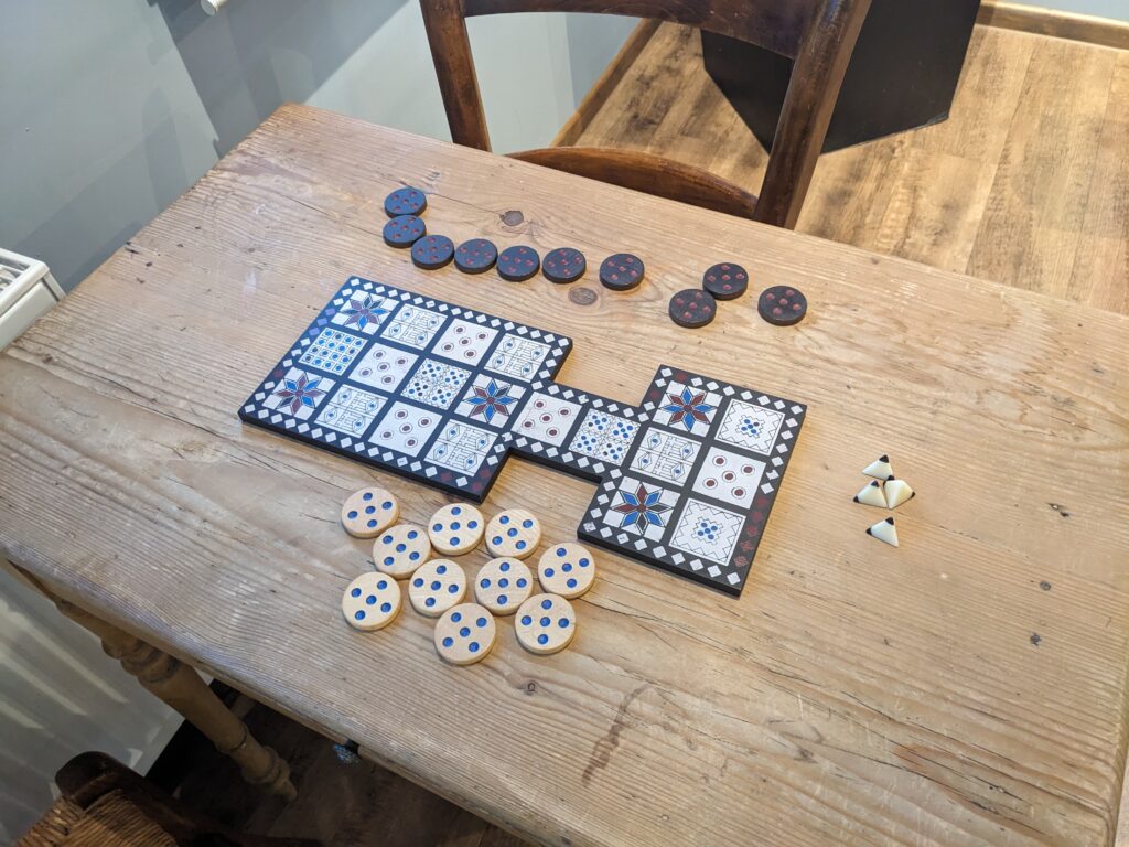 auf einem Holztisch liegt ein Spielbrett, jeweils zehn schwarze und weiße runde Spielsteine und vier Tetraederförmige Würfel