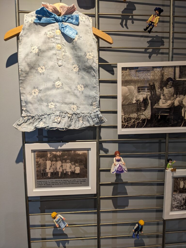 ein blaues Puppenkleid, zwei Fotos, die Kinder in typischer Kleidung vom Anfang des 20. Jahrhunderts zeigen, fünf Playmobil-Figuren