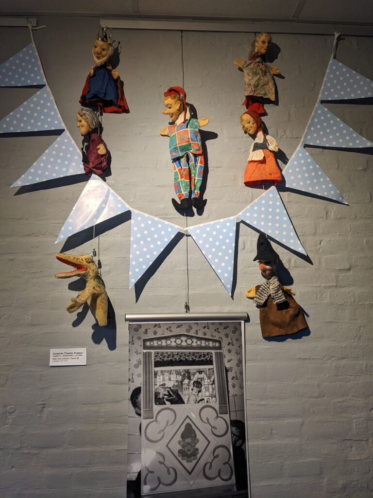 An einer Wand hängen sieben Handpuppen, eine blaue Wimpelkette und ein Foto von einem Kasperletheater