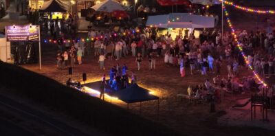 Eine große Menschenmenge versammelt sich nachts auf einem Festgelände, um eine Live-Aufführung unter bunten Lichtern zu genießen.