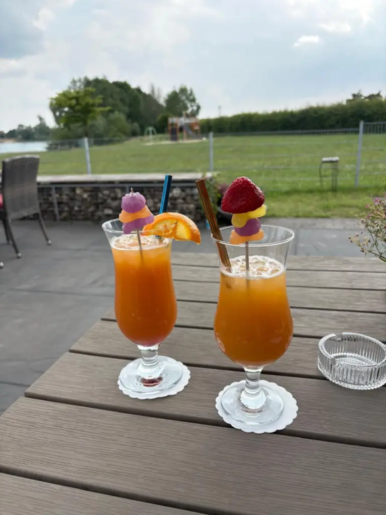 Zwei fruchtige Cocktails mit bunten Garnierungen stehen auf einem Tisch vor einer grünen Wiese und einem Spielplatz im Hintergrund.